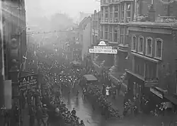 Black and white photo of people wearing military uniforms marching in close formation down a city road. Other people wearing military uniforms are standing on both sides of the road.