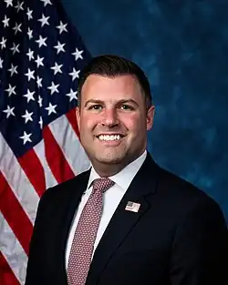 Official House portrait of Bynum smiling in front of the U.S. flag, wearing a black suit with American flag lapel pin, white shirt, and red tie with small white elephants.