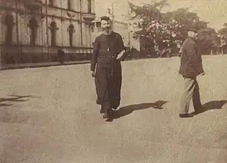 A faded snapshot of Carr Smith walking down a wide street wearing a black cassock and cap.