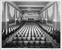 Black-and-white photo of the interior of the Revue Theatre, taken from front of room facing towards projectors, showing art deco light fixtures, and narrow wooden chairs in rows with two aisles.