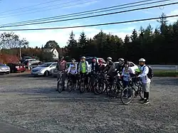 A photograph of twelve people standing in a row wearing bicycles and straddling or standing next to bicycles, all looking left under a blue sky