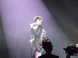 Photograph of a woman who sings on stage. She wears a dark leather blouse and a short skirt, and light tights.