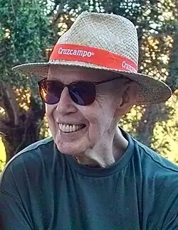 Photo of smiling older caucasian male, wearing a straw hat with red hatband, sunglasses, and a dark turquoise colored crew neck shirt.