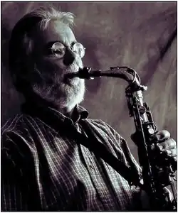 A photograph of a man with matching white hair and a beard, wearing a checkered shirt, glasses and a strap across his chest, playing the saxophone.