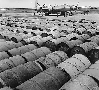 Black and white photograph of at least eight rows of fuel barrels in the foreground with a plane in the background