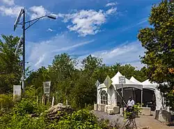 A dead end road at left from which a tall metal pole with many devices attached rises in an area surrounded by low trees under a blue sky with clouds. At lower right a dirt pathway leads a short distance to a white tent with peaked roofs next to several road signs facing the other way and concrete blocks on the ground. The one visible sign says in French and English that pedestrians are not permitted.