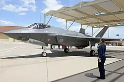 Colour photo of a grey military aircraft on the ground. A man wearing blue is standing nearby and saluting the aircraft.