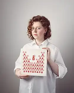 Rufina Bazlova is depicted standing. She is holding a square artwork with red pictograms on a white background. Bazlova's red curly hair is half-length and she is wearing a loose white blouse. She looks directly into the camera.