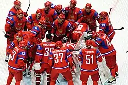 A group of hockey players stand on the ice in a circle, huddling together in front of the goal. They are all wearing red, blue and white sweaters.