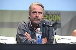 Jeremy Irons seated at a table in front of a microphone with his arms crossed