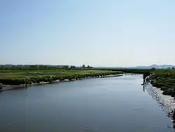 A photograph of a calm river in the middle of rural farmlands, during the day