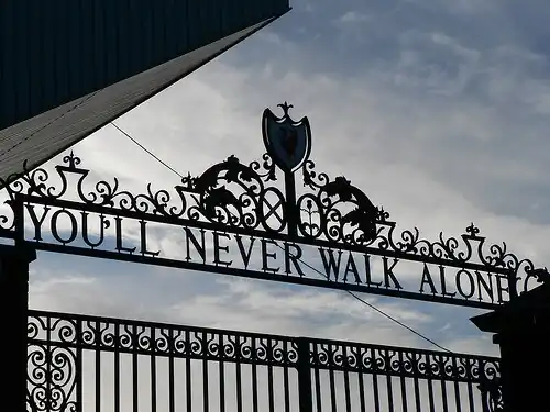 Design of the top of a set of gates, with the sky visible. The inscription on the gates reads "You'll Never Walk Alone".