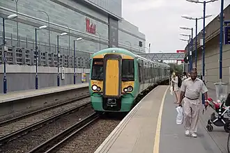 A green Southern Railways train is stopped at a platform. In the background is a silver building. A sign on the building reads, "Westfield".