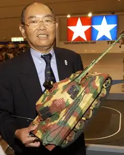 A smiling man wearing glasses and a business suit. He has a balding head and is holding a plastic model tank in his hands.