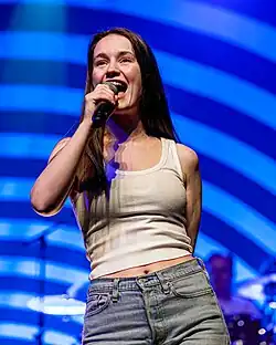 Sigrid wearing a white tanktop and blue jeans. She is singing into a microphone.