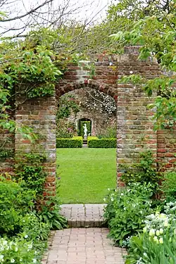 a view of an arch in a pink brick wall with a path leading to a statue
