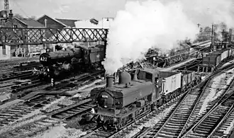 Two steam engines are passing through a station. On the left is a large passenger express locomotive, and on the right, slightly ahead, is a pannier tank locomotive pulling a goods train of open wagons and a flat bogey. It looks as though the locomotives are racing, but as the plumes of steam from the locomotives are at different angles, the fast express will probably quickly catch up with the pannier and pass it.