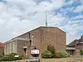A bulky, plain brown brick building. A flat-roofed projection on the near side has four evenly-spaced ground-to-roof windows with concrete details; the main part of the building behind it has evenly-spaced windows in concrete surrounds, the rightmost of which is much taller than the others. A tall, very thin green metal spire sits on the right side of the roof.