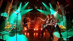 The band Speed limit playing on a brightly teal lit stage with the bass-player on the left, two guitarists on the right in the foreground and drums in the back.