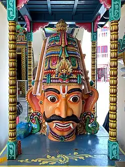 A crowned male wooden head with big eyes and ears, a Vaishnava tilak and bushy brows and moustache. A garland is seen around the neck and on the crown. He has pink skin, and two large canine teeth that hang down lower than his bottom lip. His eyes are wide open and he has large S-shaped ears.