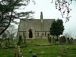A small stone church seen from the south, with an extensive roof, small lancet windows, a prominent gabled porch, and a central bellcote
