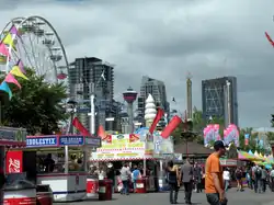 Crowds of people wander around booths selling carnival food. A merry-go-round is in the foreground to the left, and several skyscrapers stand in the background.