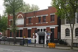 A large red-brick building with black window frames as seen from across the road with two trees in front. A sign on top of the doorway reads "Essex Road station"