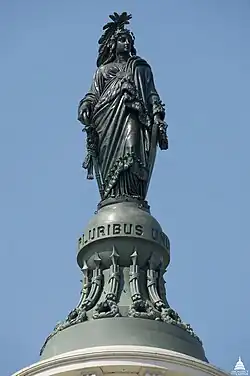 A large green bronze statue of a woman with a robe. She is holding several things, and below her are the words "E PLURIBUS UNUM".