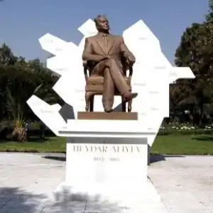 Photograph of the Heydar Aliyev statue, a bronze sculpture mounted on a marble pedestal. Aliyev wears a suit and is sat on a chair with his right leg crossed. The pedestal is inscribed with "Heydar Aliyev 1923 - 2003" in capital golden letters. Behind the statue, there is a sculptural representation of Azerbaijan featuring the Spanish-language names of several Azerbaijani cities.