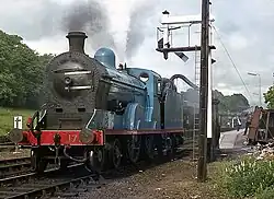 A blue steam locomotive stopped under a waterspout, currently with no train behind her. A semaphore signal hangs over the track above her, and the station platform is visible behind her, with several on-lookers.