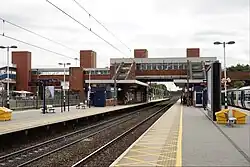 a view from a railway island platform, looking at the footbridge at the station. There is a train stopped on the right-hand side of the island platform, and overhead line equipment visible above the two visible tracks. The footbridge is made of four pillar shafts with tubular sections in between.