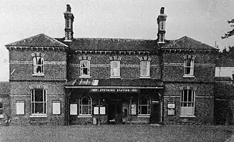 A black and white photo of a medium-sized brick building as taken from across the street. A canopy extends from the first floor with a white sign reading "STEYNING STATION"