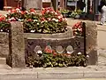 Stocks, made from central dark wooden stock beams with two holes in. The beams are supported by stone pillars. They have flowers planted in front of them, and the market cross, also with flowers, sits behind.