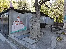 A miniature stone memorial tower, resembling a traditional Svan tower from Georgia, stands on a concrete platform in an outdoor common area. Behind it is a gray wall with a colorful mural of a pink church with a red roof. To the right are two wooden benches, and a large tree grows behind the memorial, under a canopy of other leafy trees.