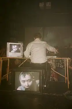 Sweatmother faces away from the camera, wearing a white shirt and black leather trousers. He is surrounded by monitors, and controls a visual synthesiser to a crowd of people at a rave.