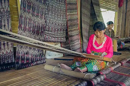 T'boli dream weavers using two-bar bamboo backstrap looms (legogong) to weave t'nalak cloth from abacá fiber. One bar is attached to the ceiling of the traditional T'boli longhouse, while the other is attached to the lower back. The cloth is being patterned by dying the warp, so the loom equipment is simple; a heddle rod, a shedding stick, and a batten. She is also using a footrest. Philippines.[43][44]