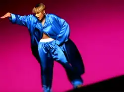 A women showing a white boxer inside her pants, with her right hand up and her left hand on her hip, backed by a dark pink background.