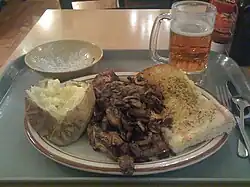 Plate with a baked potato, steak topped with mushrooms, and a piece of garlic toast. This is on a cafeteria-style tray with an empty salad bowl and a mug of beer.