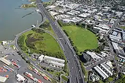Overhead view of a motorway running through a circular green park, with water to the top left and industrial buildings elsewhere
