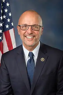 A chest-up photo of bald man wearing glasses and a suit, in front of a blue background