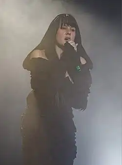 Teya Dora wearing a black outfit, singing onstage, surrounded by smoke