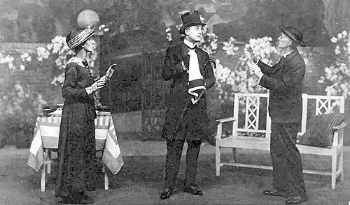 stage scene in a garden setting with a man in full mourning costume centre, older woman to his right and an older man in clerical garb to his left, all wearing hats