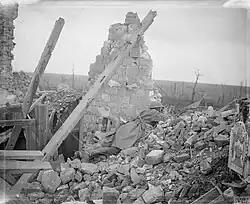 Black and white photograph of a destroyed house with one remaining wall and visible roof timbers. Image of soldier wearing a Brodie helmet and great-coat with a rifle lying prone, peering over rubble towards the top right of picture. In the background are mangled tree trunks, stripped of their foliage.
