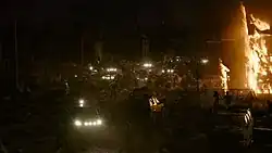 A cul-de-sac is overrun by dozens of infected creatures, included a very large one near the center. A building to the right is on fire.