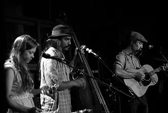 A black-and-white image of three musicians (a woman and two men) playing instruments.