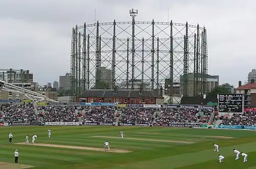A cricket match in progress; gasholders are visible in the background
