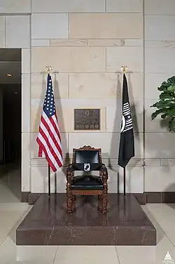 A black wood-and-leather chair. On the right is a POW/MIA flag, and on the left is an American flag. Behind the chair is a plaque with unreadable text.