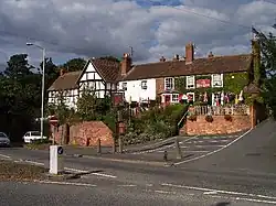 Red brick and tile building on a sloping site with outside raised seating area and car parking to front at lower level adjacent to road down on a sunny day