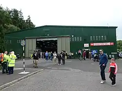 Green hangar structure with red-on-white signs advertising the 'Exhibition Hall' and its shop and cafe.