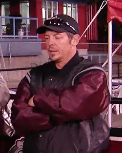 Half-length view of a person in his early 40s. He is standing upright with his arms folded across his chest. He is wearing a black and maroon coat and a baseball cap.